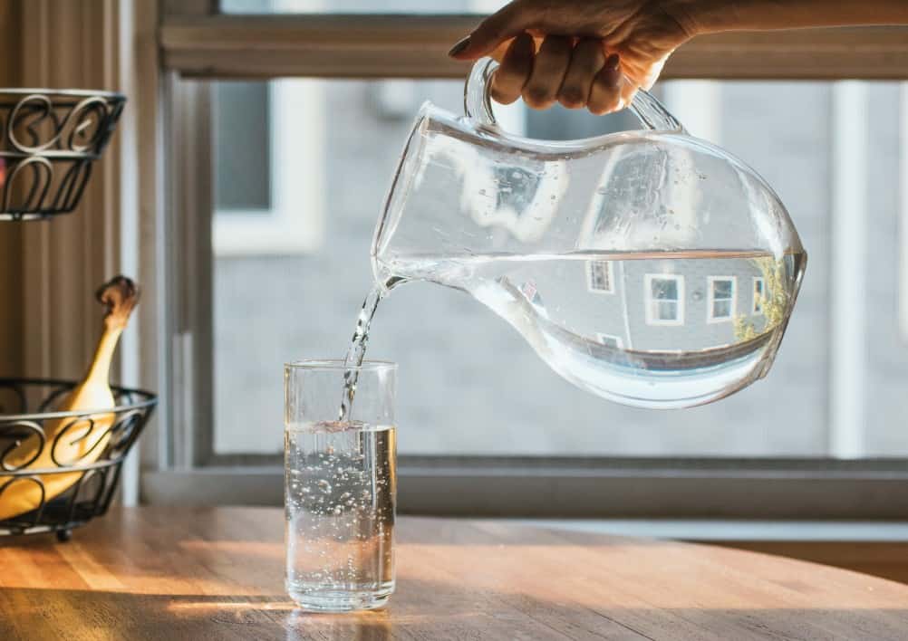 Pouring a glass of water out of a glass pitcher into a clear glass sitting on a wooden countertop with a banana in the background